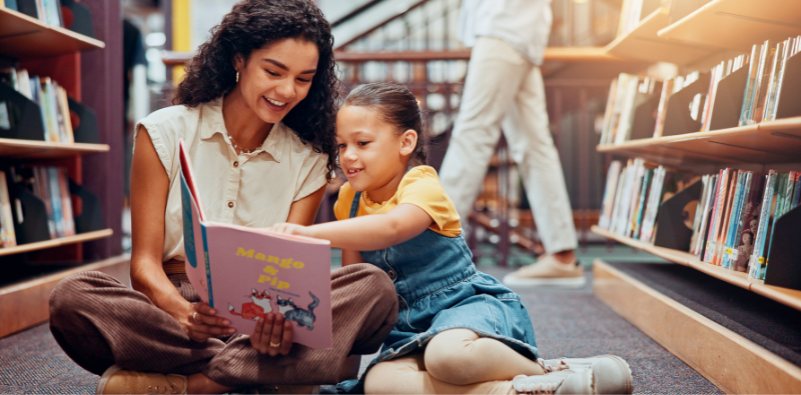 A parent and child reading a book sat on the floor in a library.