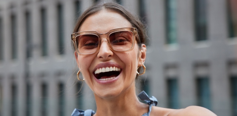 A smiling, middle-aged woman, wearing a pair of transparent sunglasses outdoors