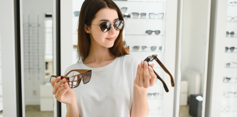 A woman at the opticians wearing a pair of sunglasses, and holding a pair in each hand.