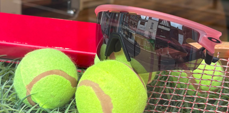 Pink Oakley sunglasses sitting on top of a tennis racket and tennis balls.