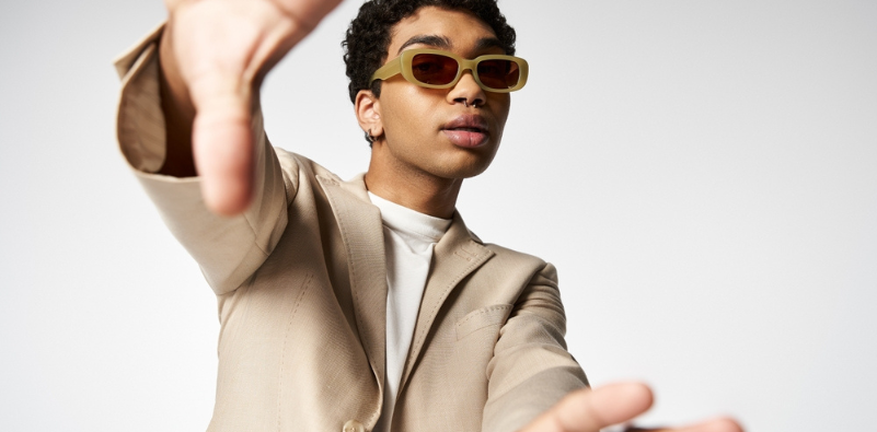 A stylish young man wearing neutral coloured rectangular sunglasses, with his hands reaching out