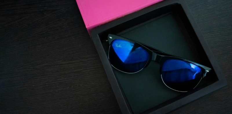 Photo by Lenzil Gonsalves on Unsplash. A bird’s eye view of a black Ray Ban original Wayfarer sunglasses frame with a blue lens tint in a presentation tray on a dark wood grain table