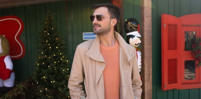 Brunette man standing outside with a Christmas tree behind him looking to his right wearing a thin winter coat with hands and brown tinted aviator sunglasses