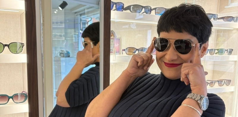 Staff member from The Optical Studio opticians posing in practice wearing a pair of aviator sunglasses smiling with her forefingers up against the side of the frame