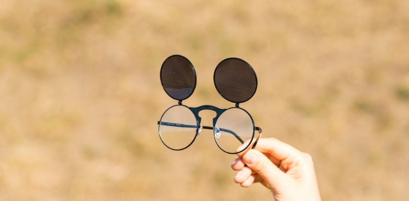 A hand holding out a pair of round glasses frames with brown tinted flip up sunglasses attached to them outside.