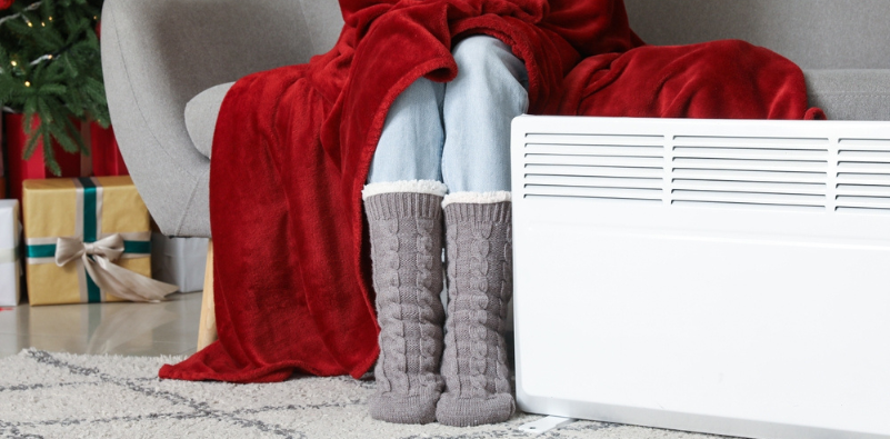 Young woman in winter socks with warm plaid near electric heater at home