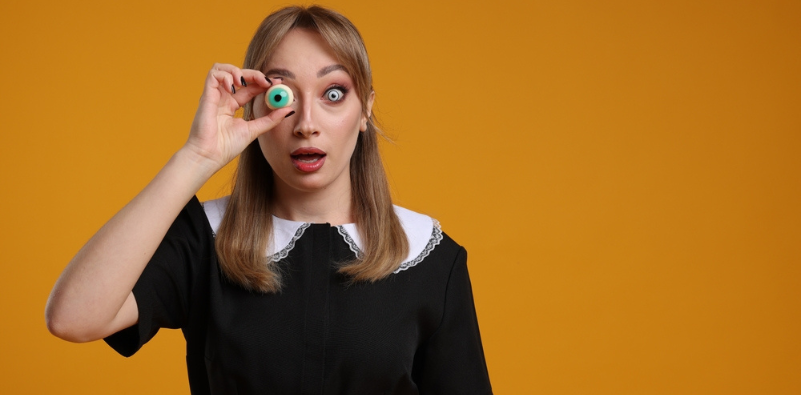 A young woman in a maid costume holding a decorative eyeball over her right eye, and a cosmetic contact lens in the other